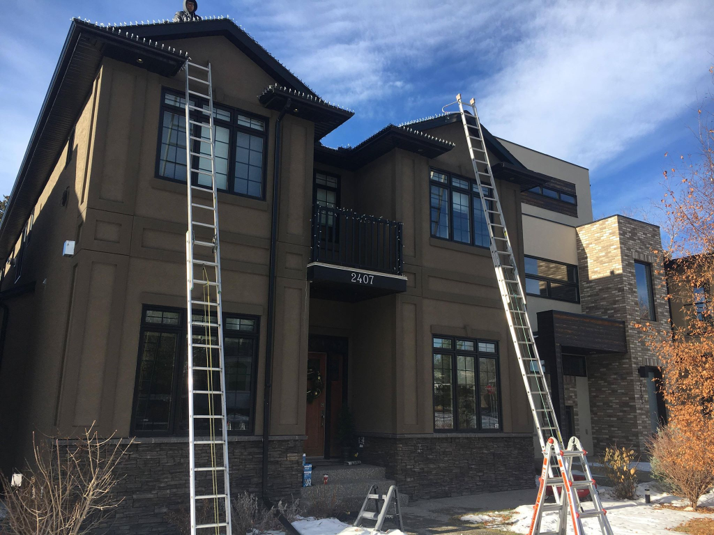 Professional Christmas light installation Calgary — ladders set up on a two-story home with a technician safely installing LED holiday lights on the roofline