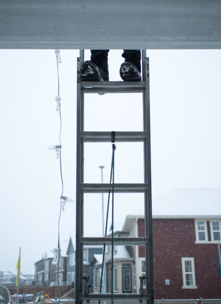 An employee standing on a ladder in snowy Calgary weather while hanging Christmas lights, illustrating the safety risks and hidden expenses often overlooked in DIY holiday lighting costs.